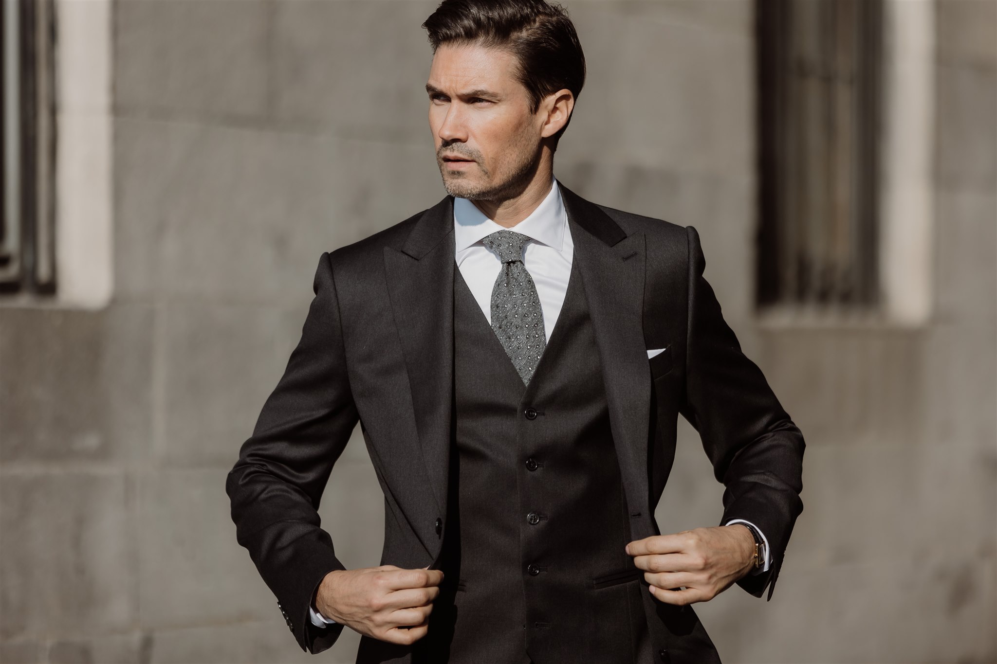 Homme élégant portant un costume trois-pièces noir avec gilet assorti, chemise blanche et cravate texturée grise, illustrant le retour du style classique et intemporel dans la mode masculine moderne.